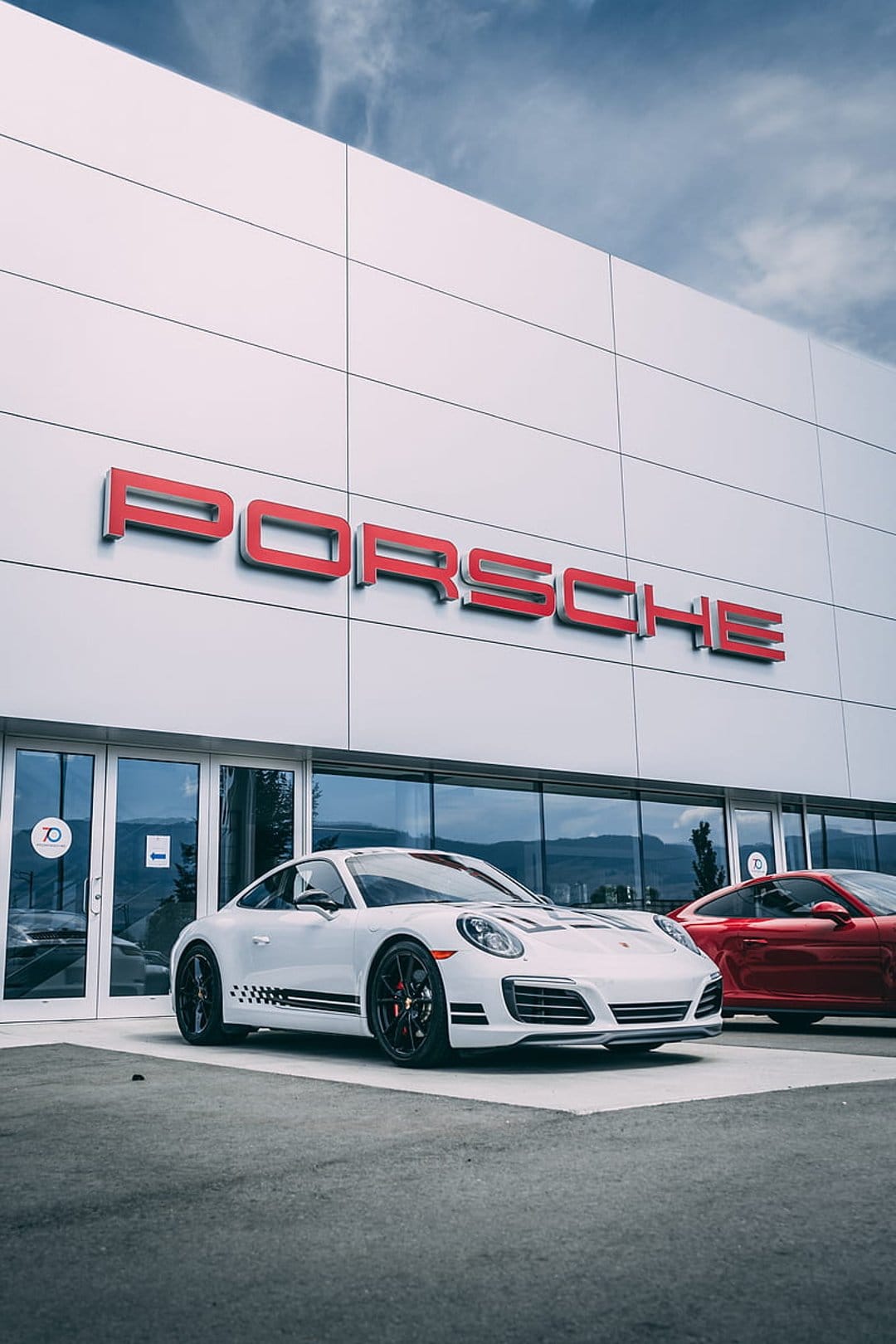 White Porsche 911 with racing stripes and a red sports car parked in front of a white luxury car dealership.