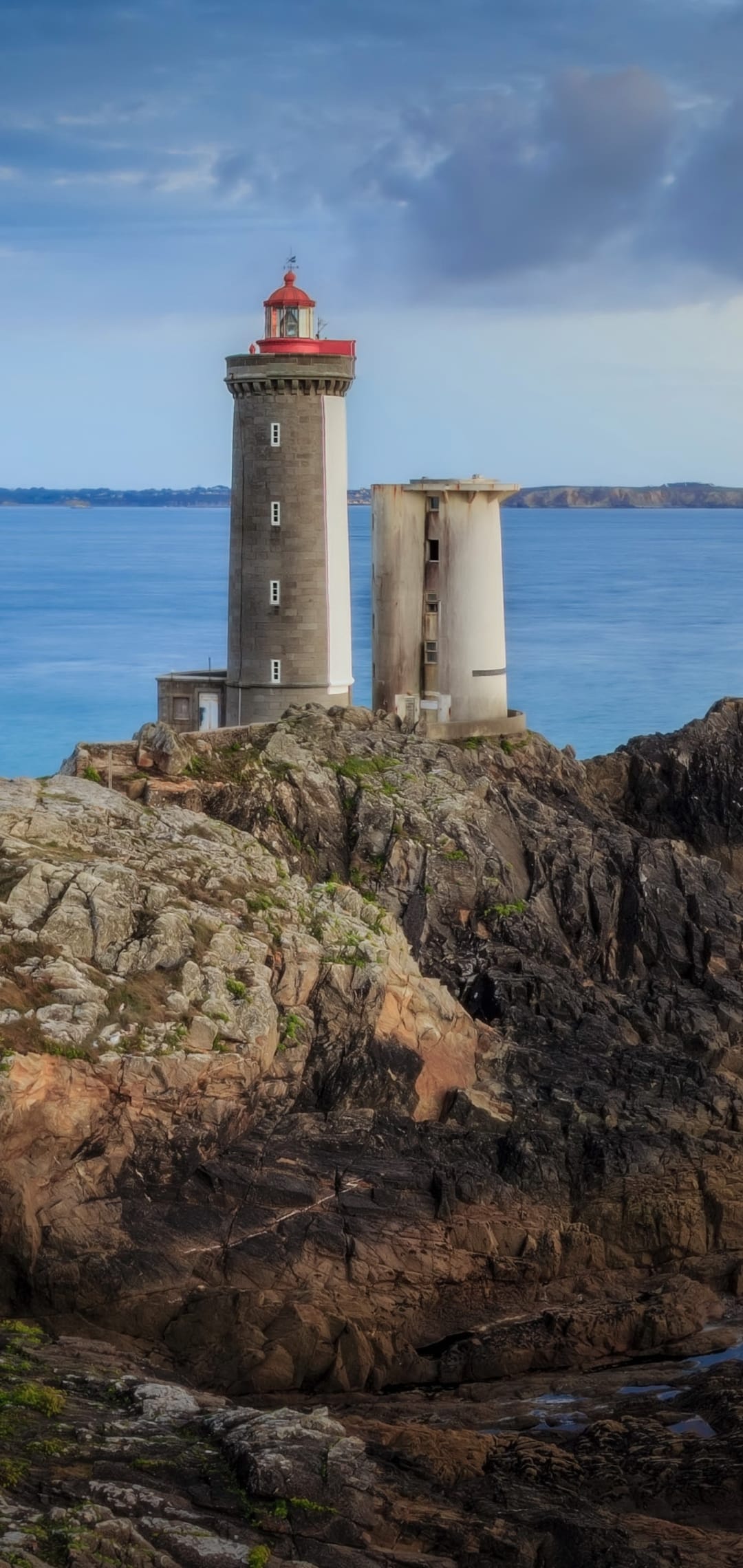White lighthouse with a red top perches on dark rocky cliffs above a deep blue sea under a cloudy sky.