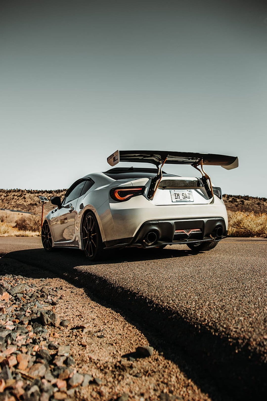 Silver Subaru BRZ with a large rear wing parked on a desert road during golden hour with a custom license plate.