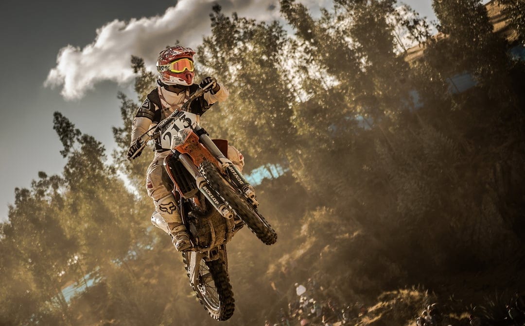 Motocross rider jumping through sunlit dust with trees and a cloudy sky in the background.