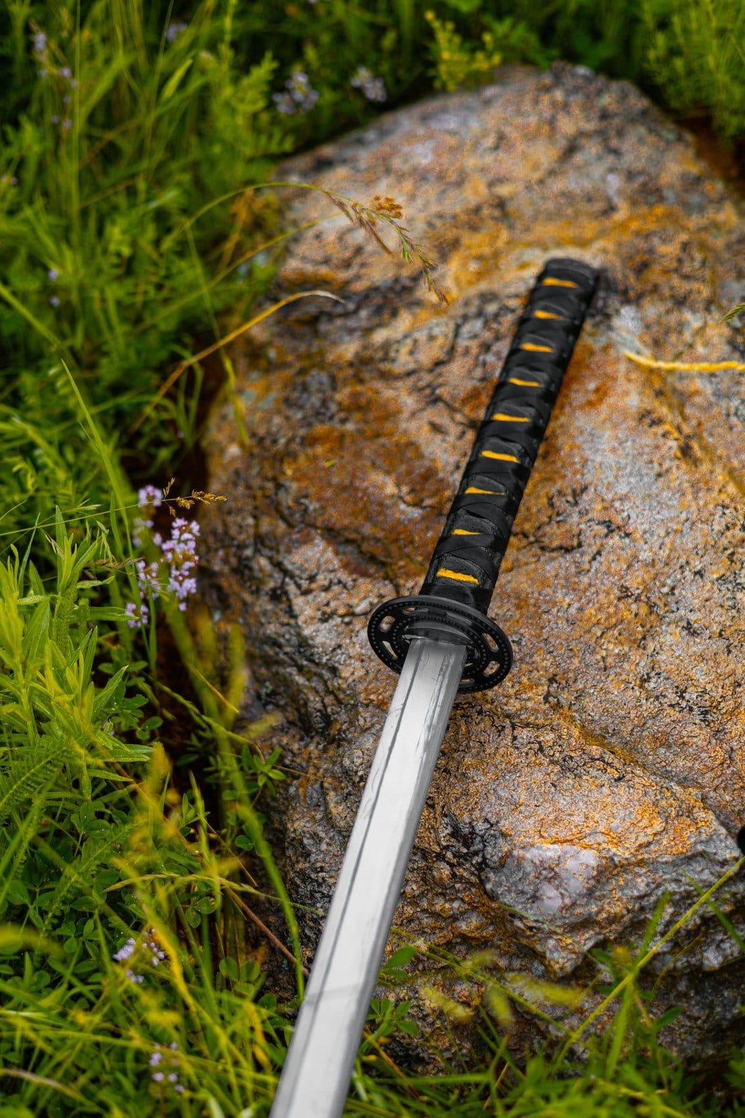 Katana sword with black and orange hilt leans against a grey rock in green grass with small purple wildflowers.