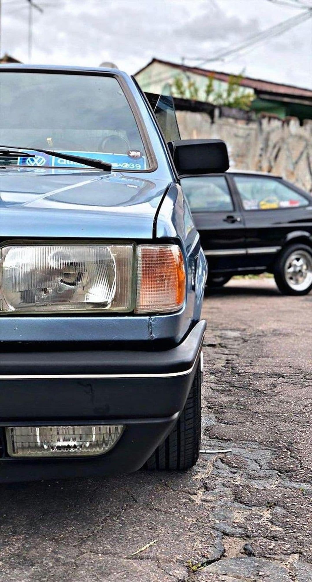 Blue car headlight and bumper on cracked asphalt with a black car and cloudy sky in the background of the shot.