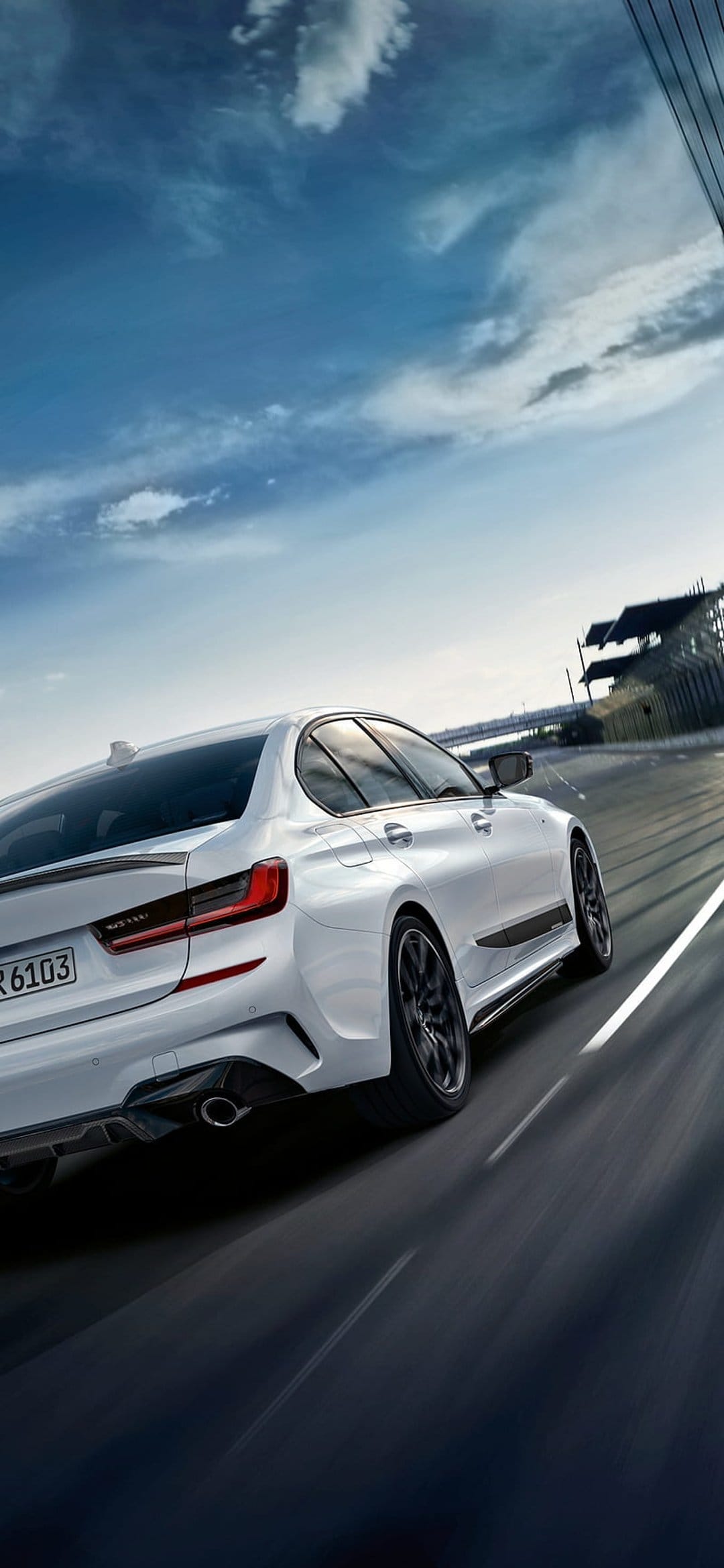 White BMW M340i sedan speeds along a paved race track under a bright sky with scattered clouds in a vertical view.
