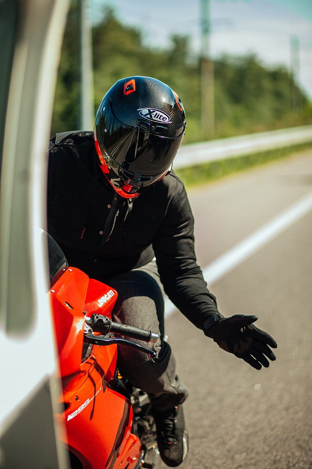 Motorcyclist in black gear sits on a red Ducati sportbike on a sunny roadside with trees under a bright blue sky.