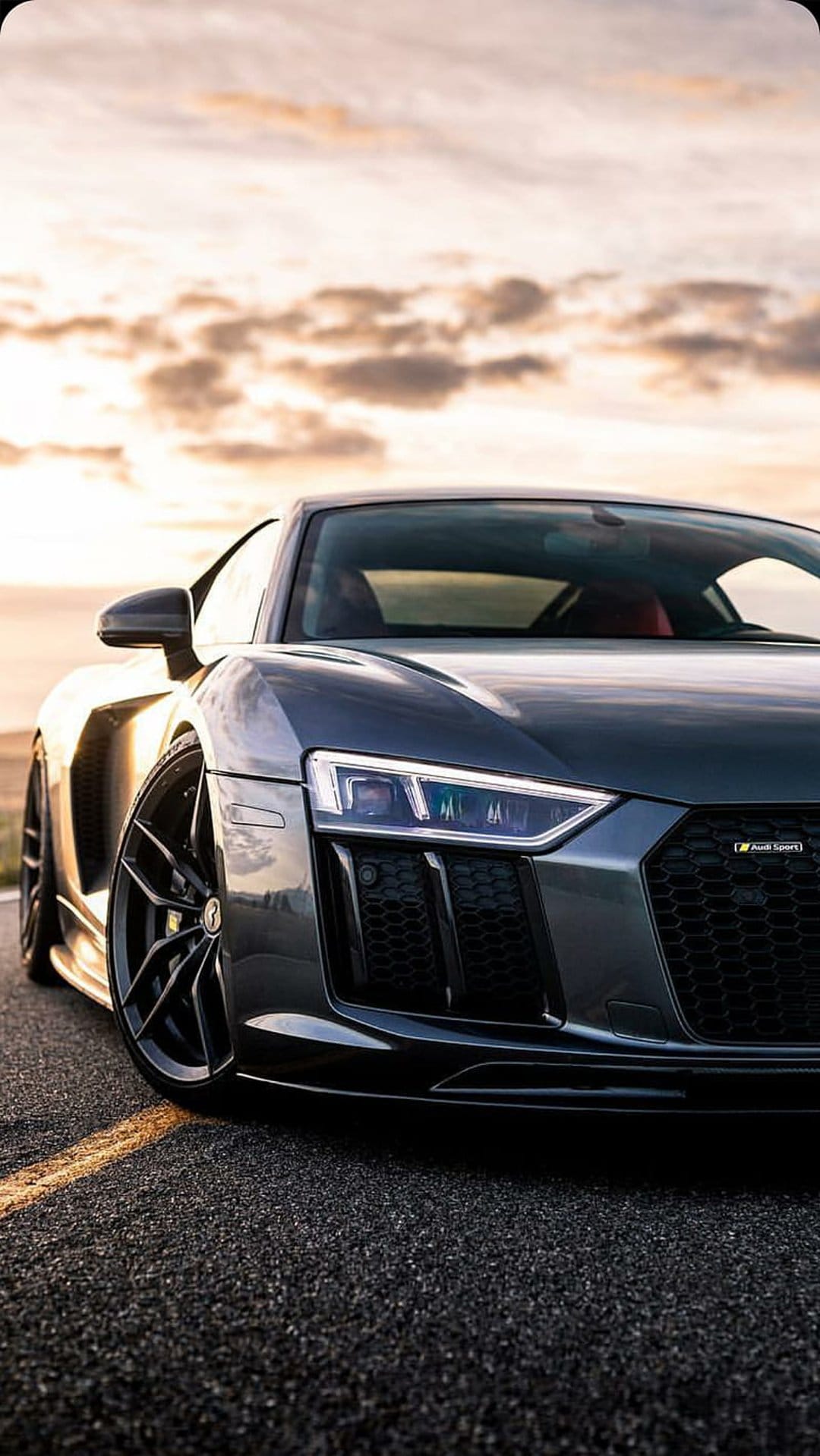 Audi R8 with glowing headlights parked on an asphalt road under a dramatic sunset sky with orange clouds.