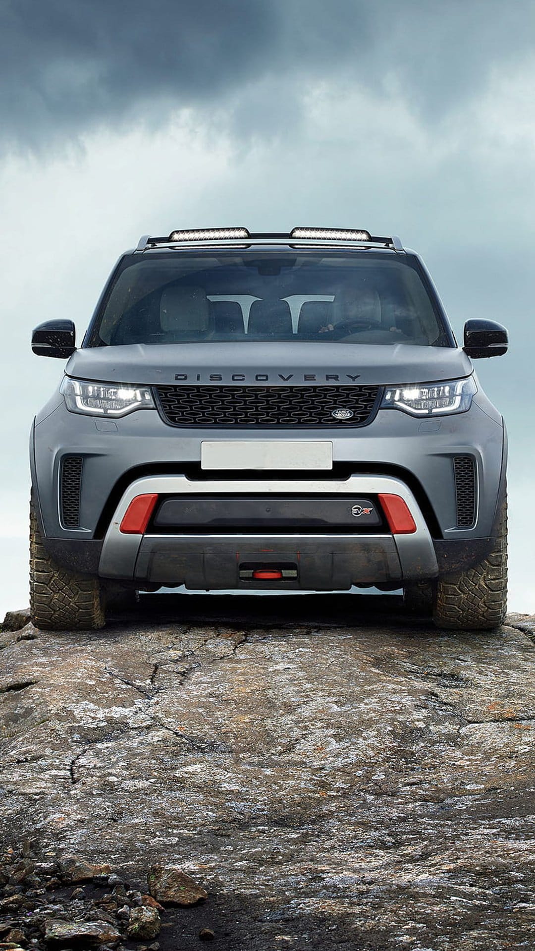 Gray Land Rover Discovery SUV with a roof rack and red bumper accents parked on a rocky outcrop under cloudy skies.