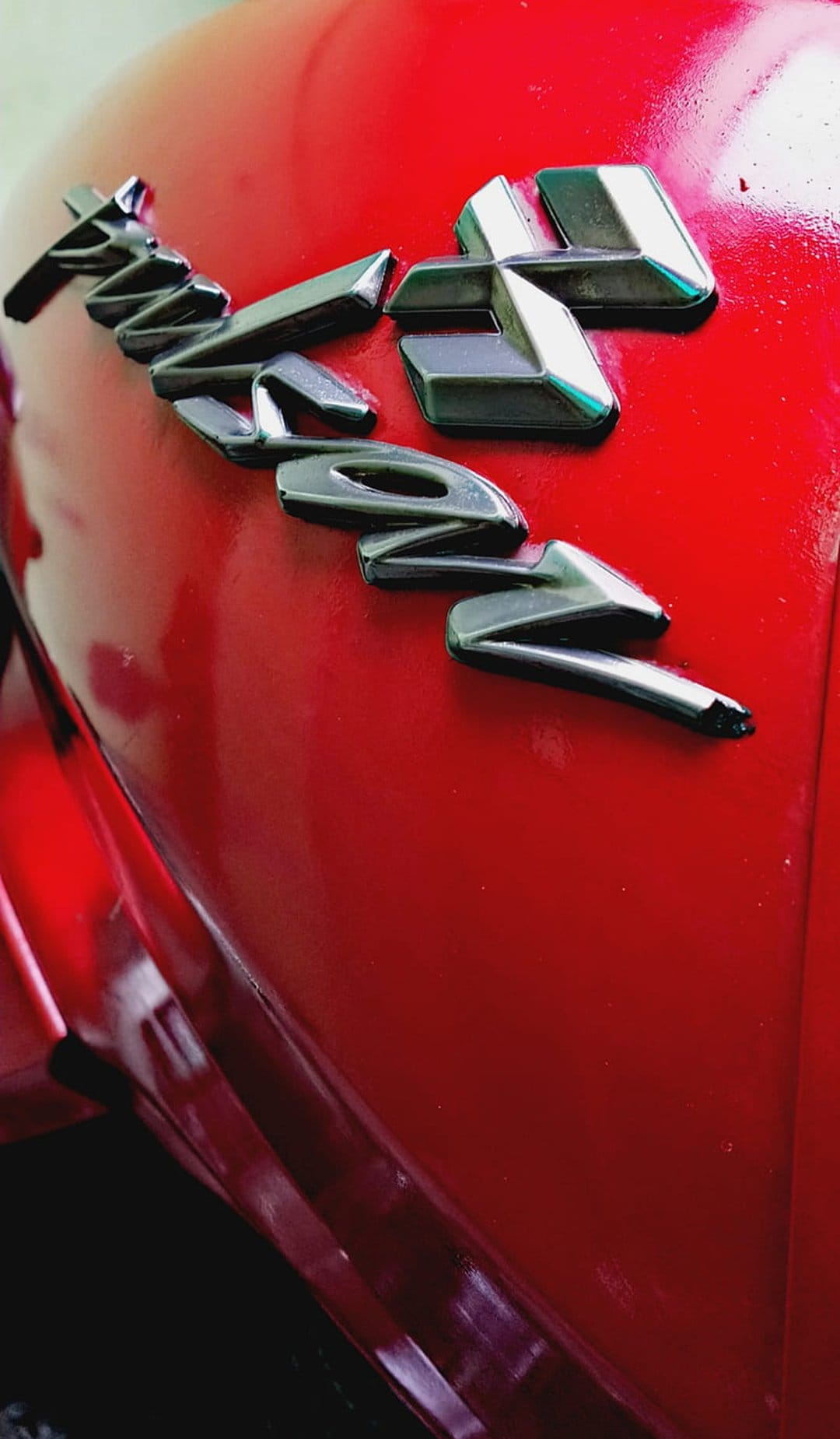 Chrome TVS emblem mounted on a glossy red motorcycle fuel tank with sharp metallic reflections and highlights.