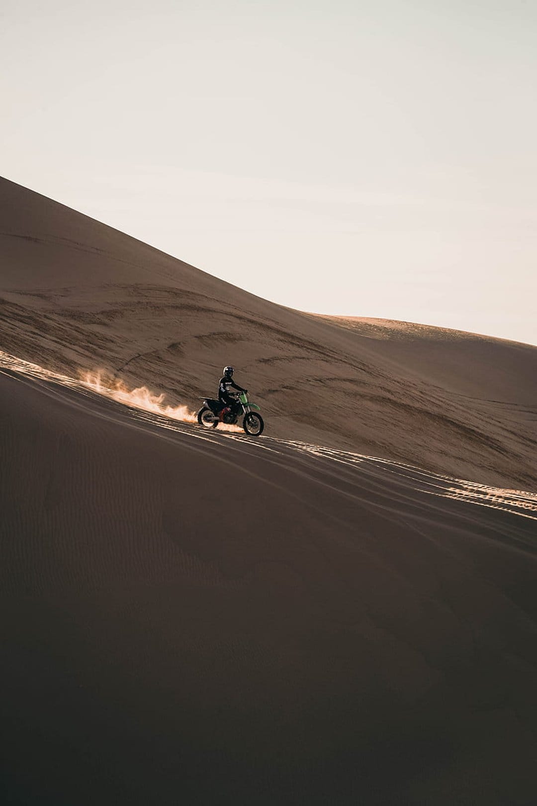Motorcyclist riding down a sand dune at sunset with golden light and a dust trail in a vast desert landscape.