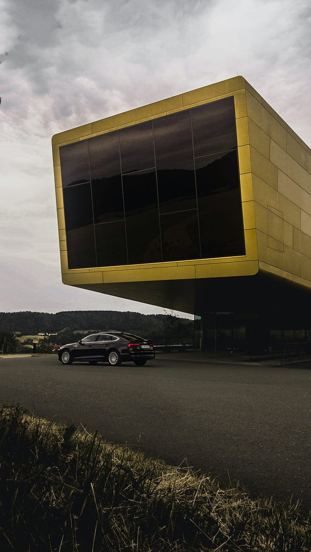 Black luxury sedan parked on a mountain road beside a geometric golden building with a dramatic overhanging roof.