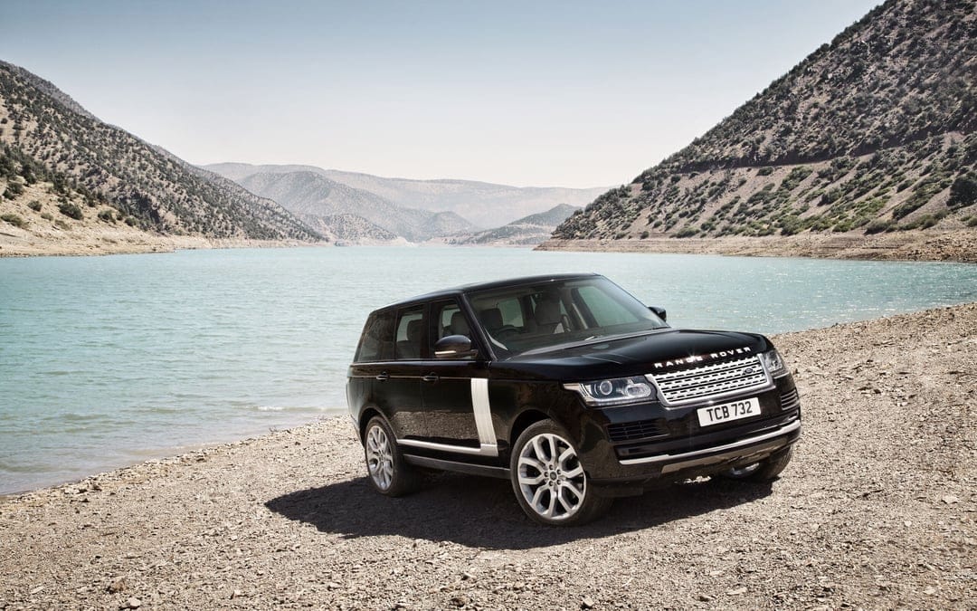 Black Range Rover SUV parked on a gravel shore beside a turquoise lake with mountains in the background.