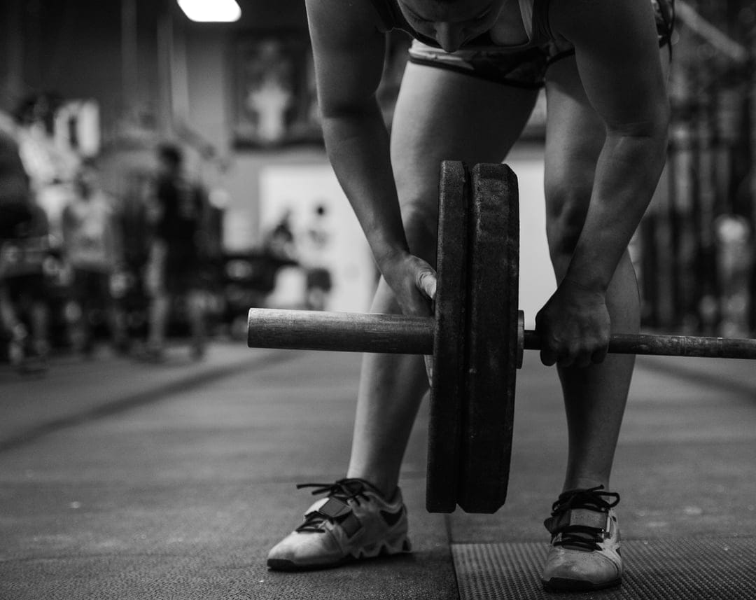 Person from the waist down grips a weighted barbell on a gym floor in a high-contrast black and white style.