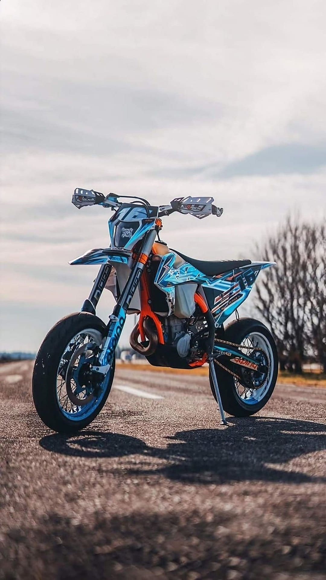 Supermoto motorcycle with blue graphics and an orange frame parked on an asphalt road under a cloudy sky.