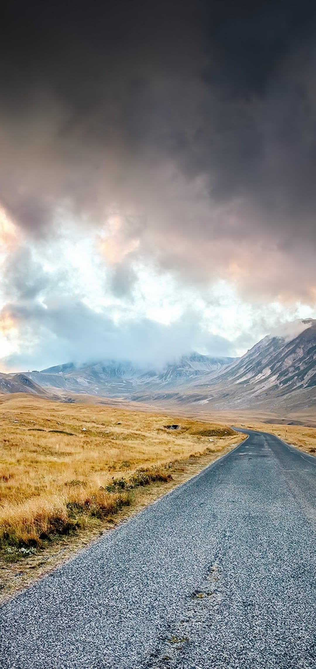Winding asphalt road through golden grasslands toward distant misty mountains under a dramatic dawn sky.