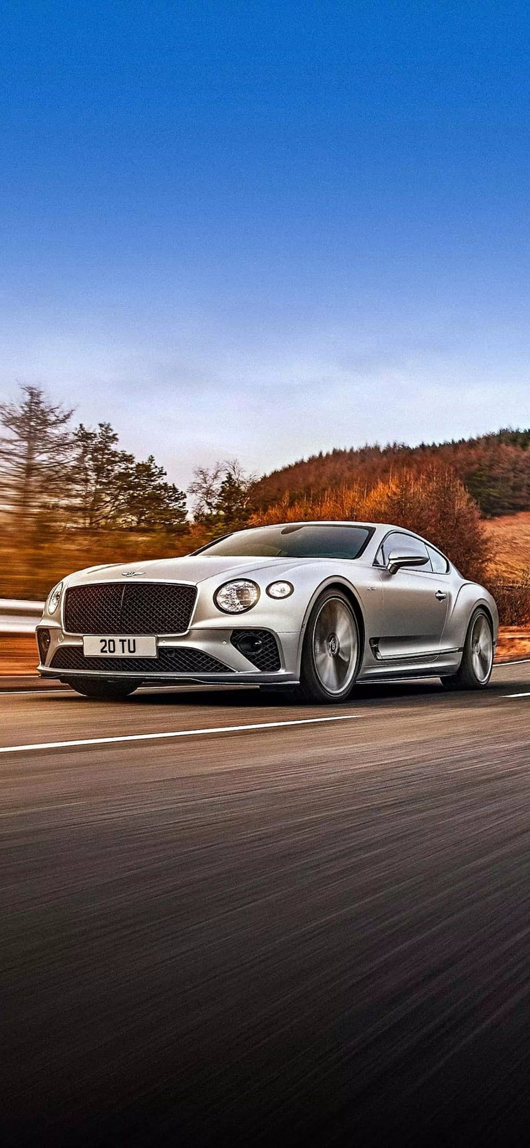 Silver Bentley Continental GT racing on a road with blurred autumn trees and a clear blue sky background.