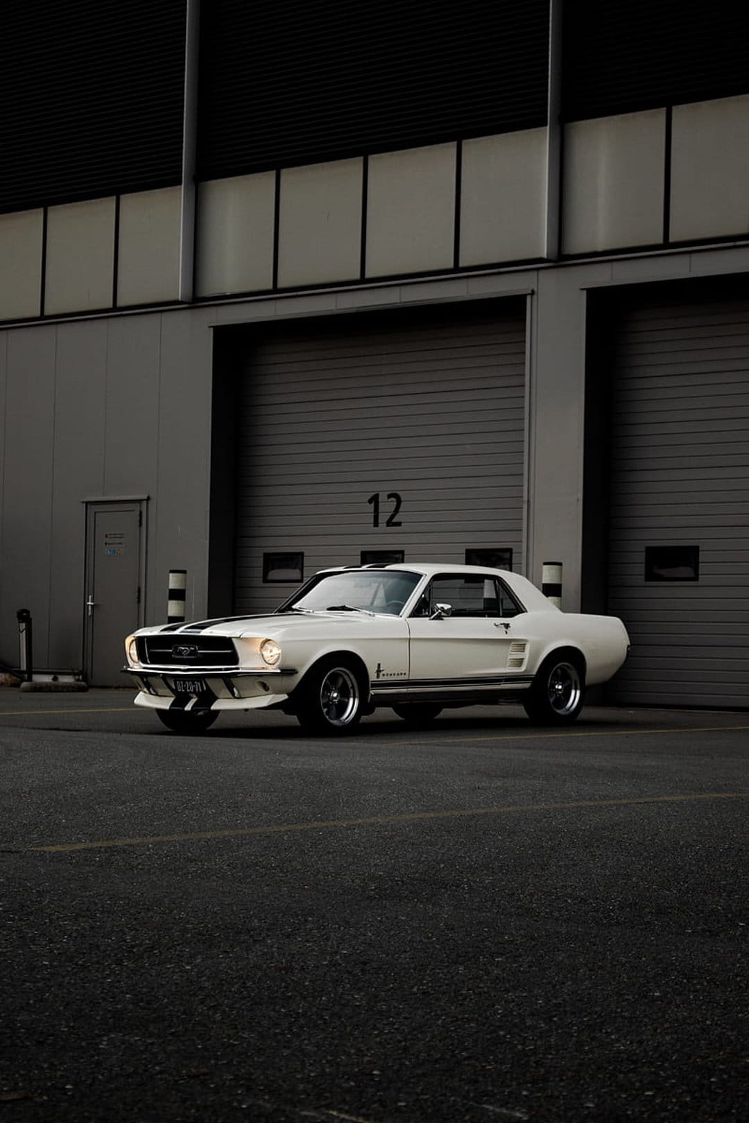 White 1960s Ford Mustang with black stripes parked on asphalt by an industrial building with garage doors.