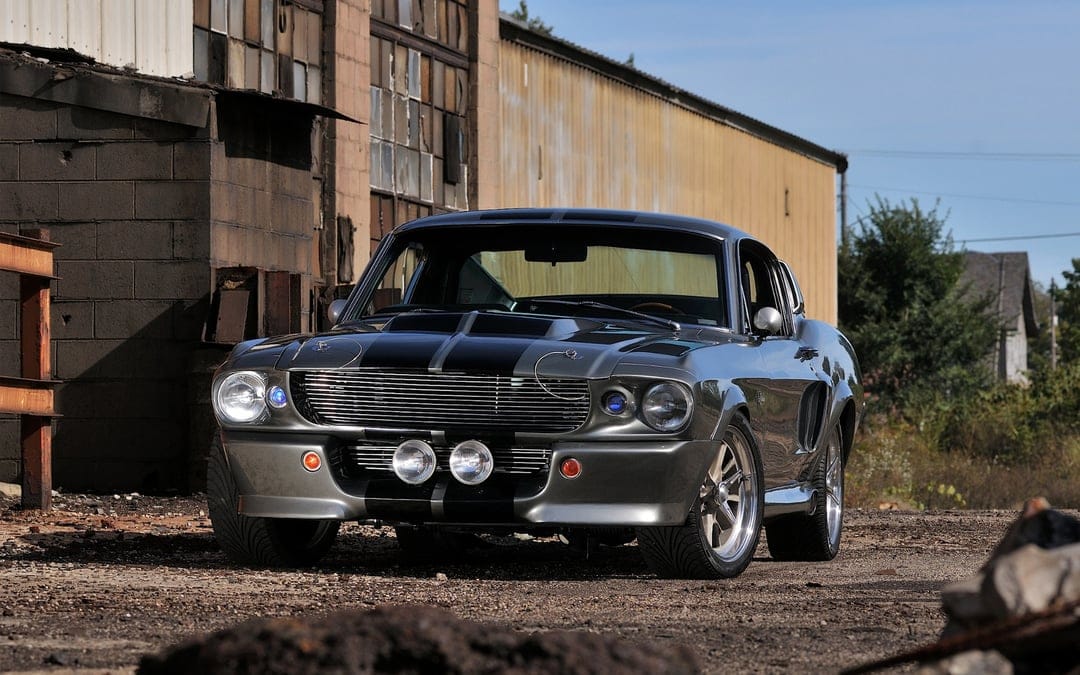 Silver Shelby GT500 Eleanor muscle car with black racing stripes parked in front of a rustic industrial building.