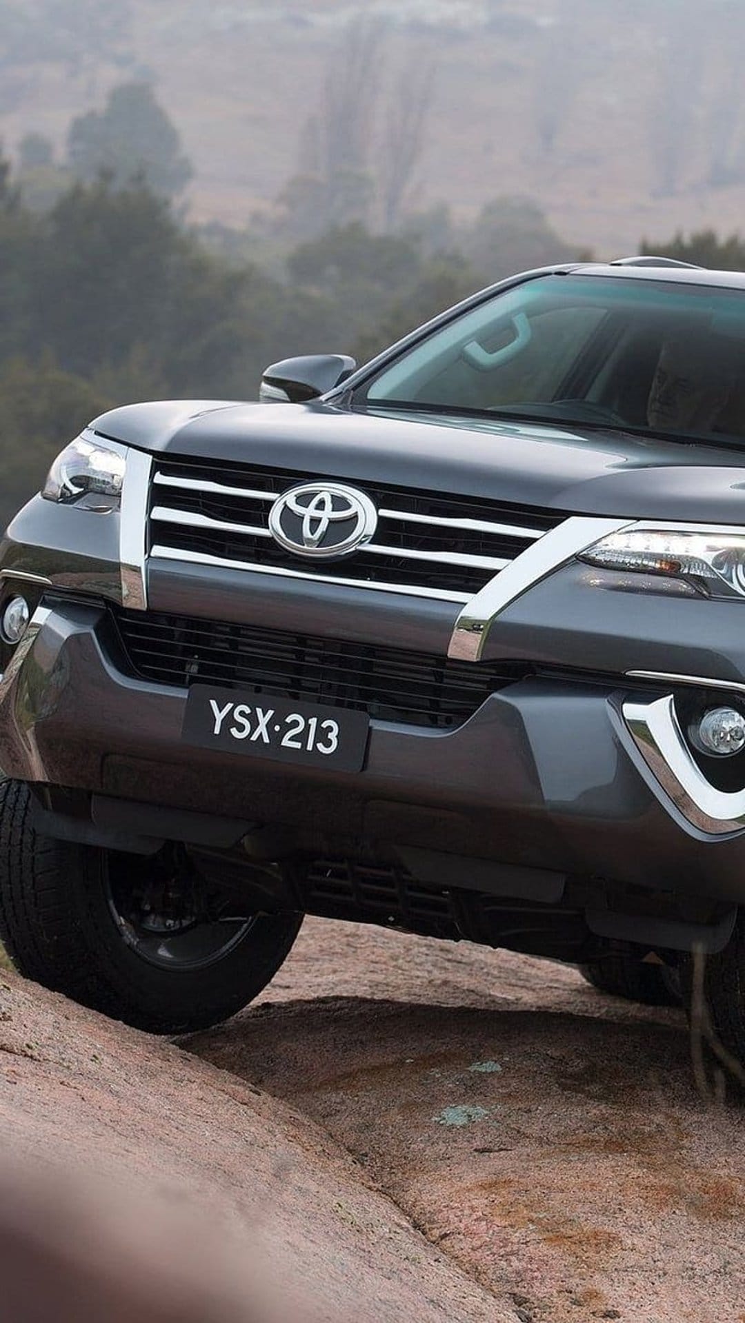 Toyota Fortuner SUV with headlights on navigates a steep rocky trail in a misty, pine-covered forest area.