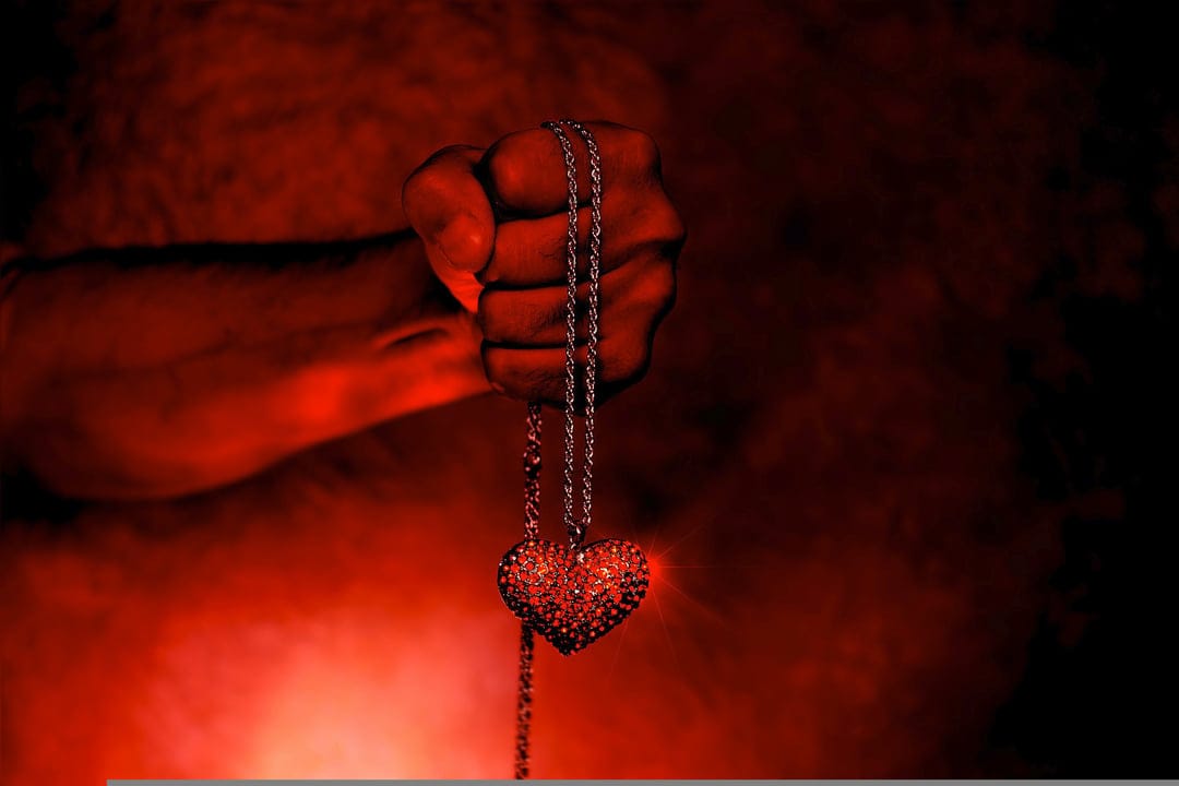 Clenched fist gripping a silver heart pendant within a dense crimson mist and atmospheric red lighting.
