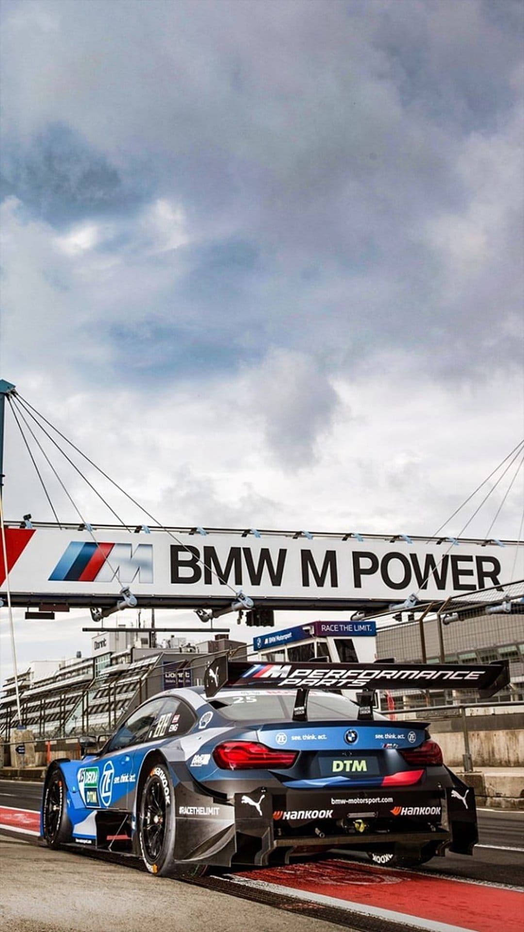 Blue and white BMW DTM race car on a track beneath a large M Power banner and a cloudy sky in pit lane.