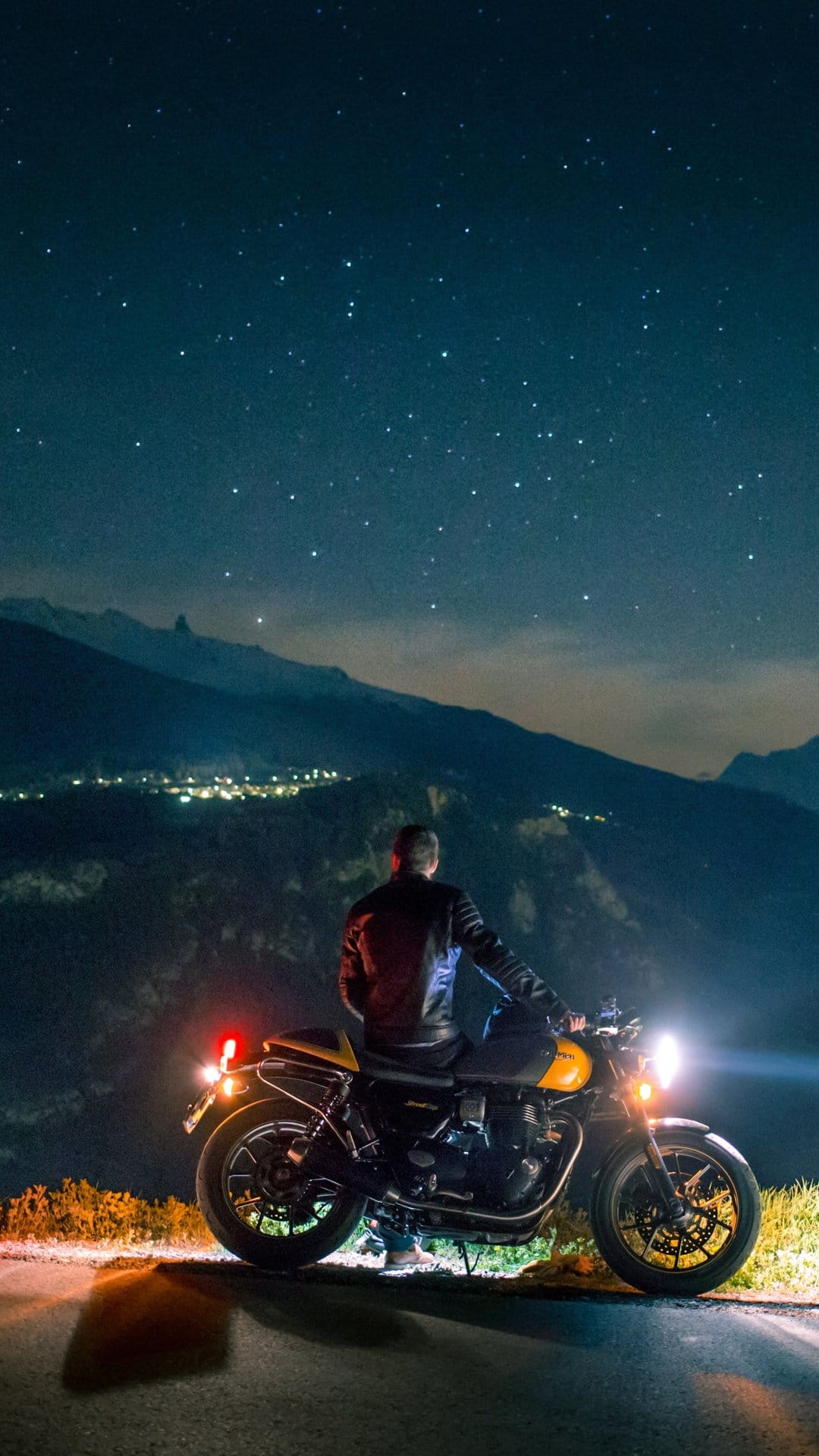 Biker on a motorcycle at night views a distant valley town under a star-filled sky with visible nebula clouds.