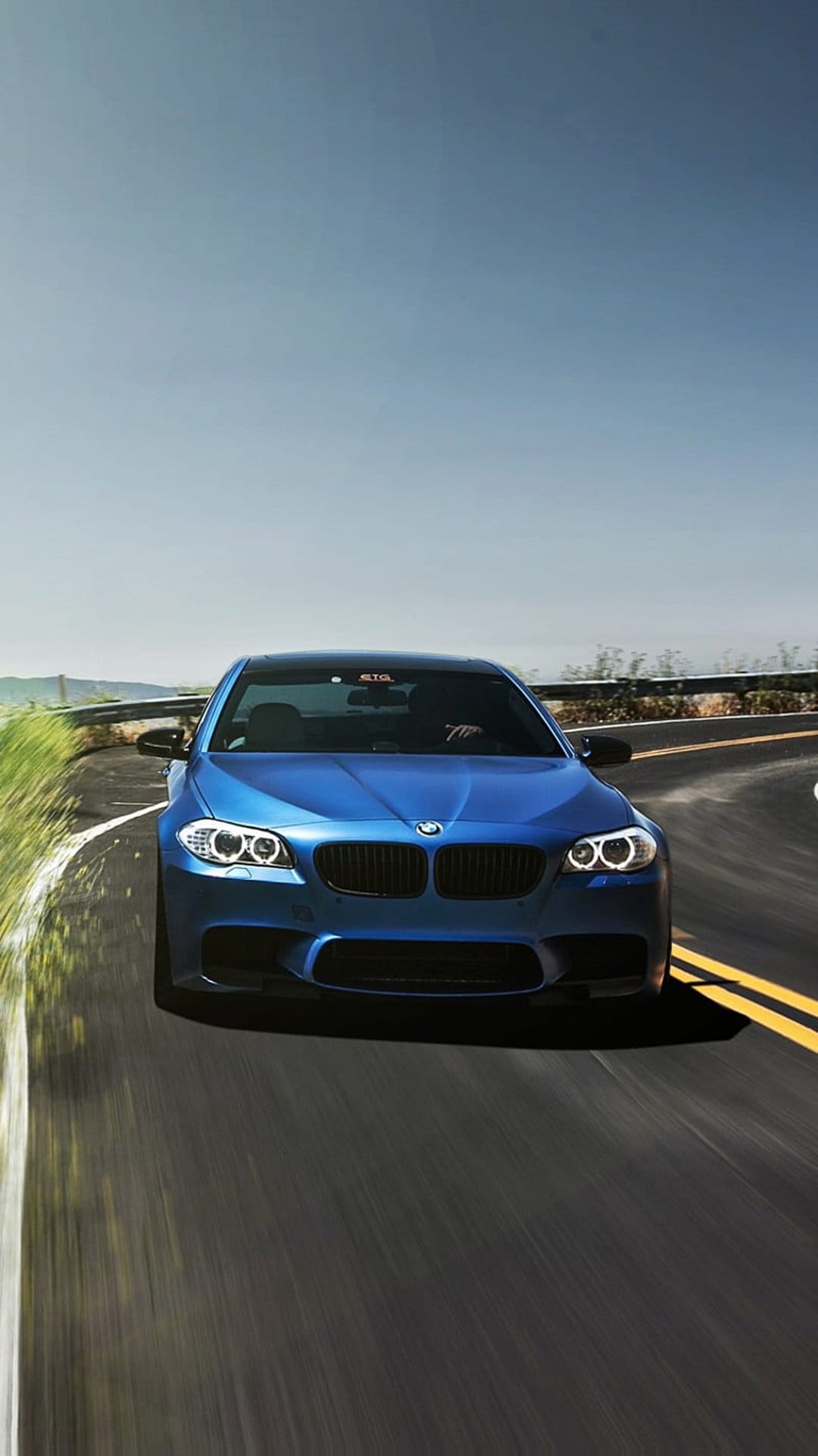Blue BMW M5 sedan drives on a winding asphalt road surrounded by green trees under a clear sky during the daytime.