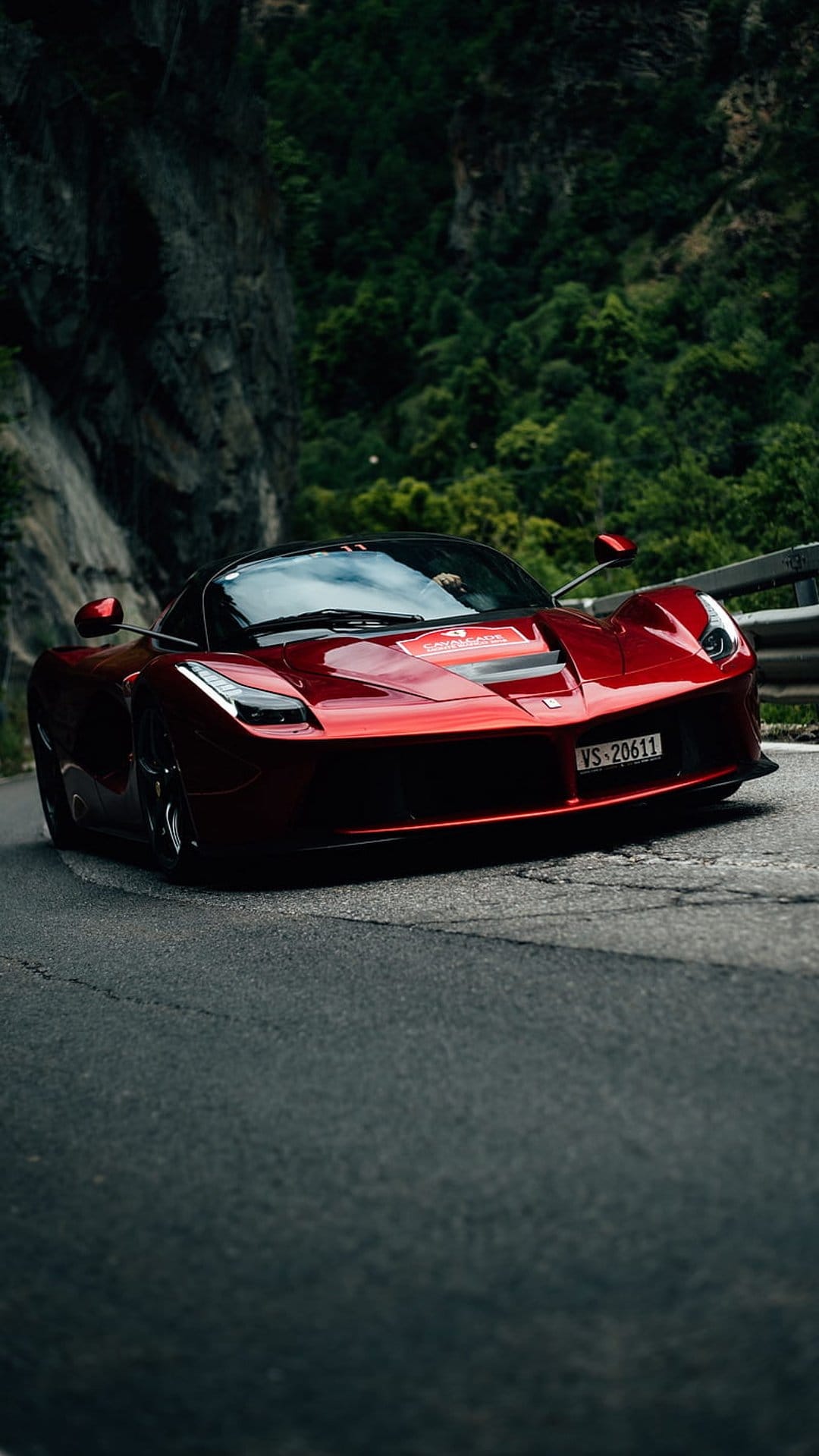 Red Ferrari LaFerrari hypercar driving on a winding asphalt road through a lush, rocky mountain landscape.