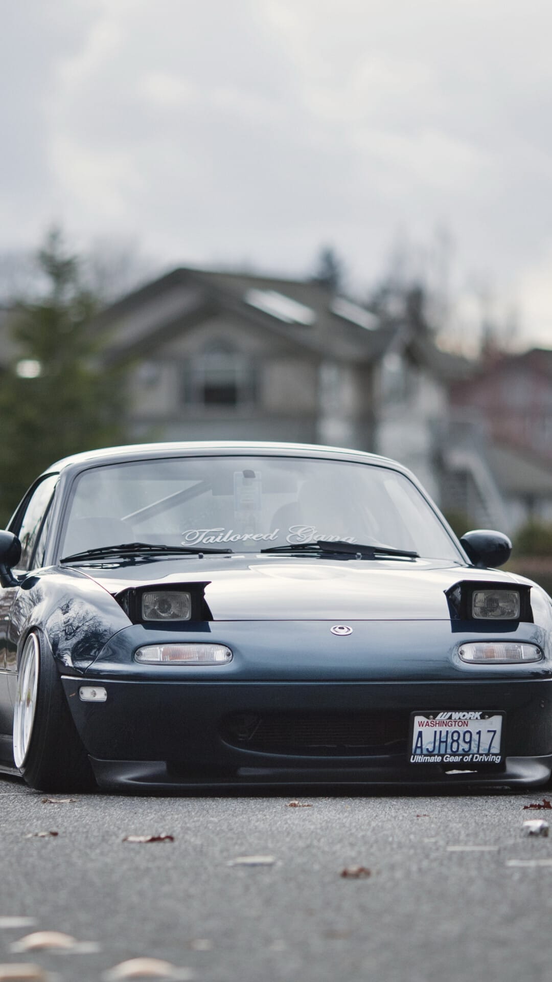 Front view of a stanced dark blue Mazda Miata with pop-up headlights and white wheels on a residential street.