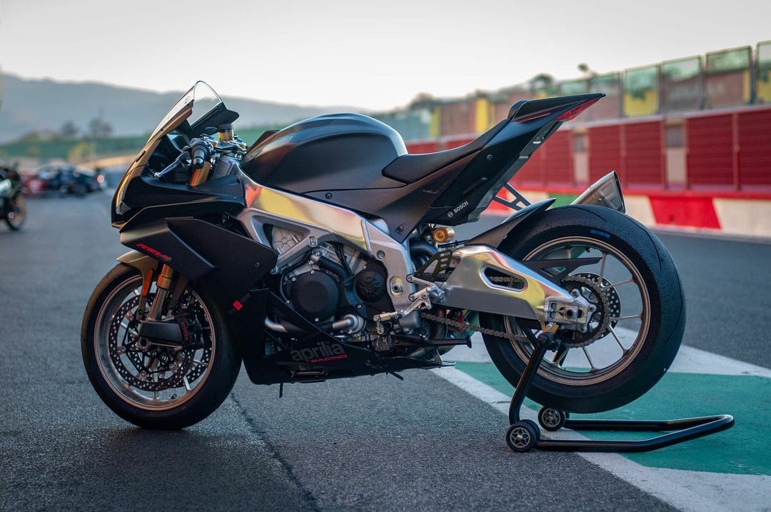Matte black Aprilia RSV4 motorcycle on a paddock stand at a racetrack during sunset with soft bokeh hills.