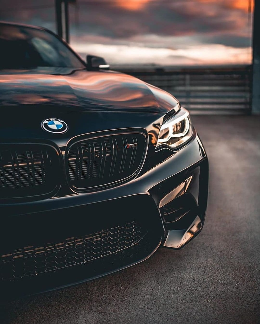 Black BMW M2 sports car with glowing LED headlights parked under a dramatic orange and purple sunset sky.