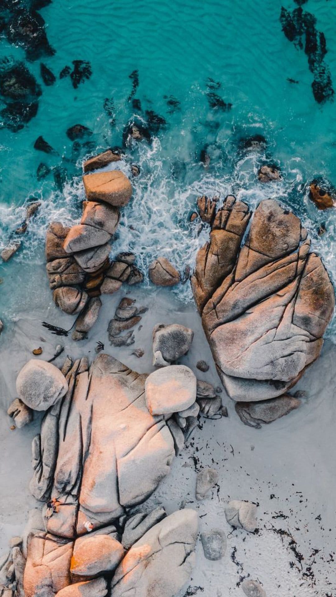 Massive sun-lit boulders sit on a sandy shore as turquoise ocean waves create white foam patterns from above.