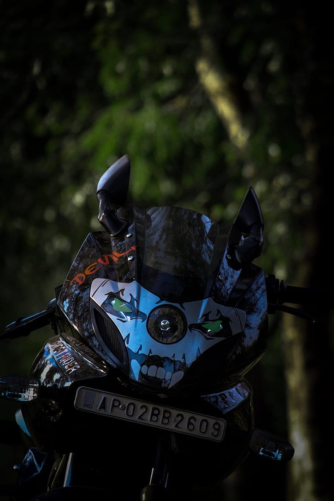 Motorcycle fairing with a skull graphic, glowing green eyes, and orange text against a dark forest background.