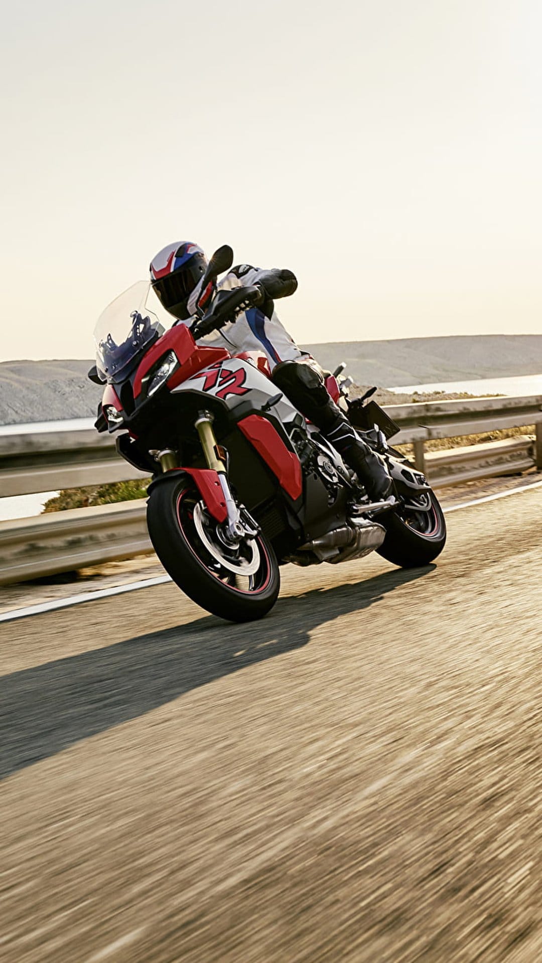 Red sport-touring motorcycle leans into a sharp asphalt turn with a calm blue lake and hazy shore in the background.