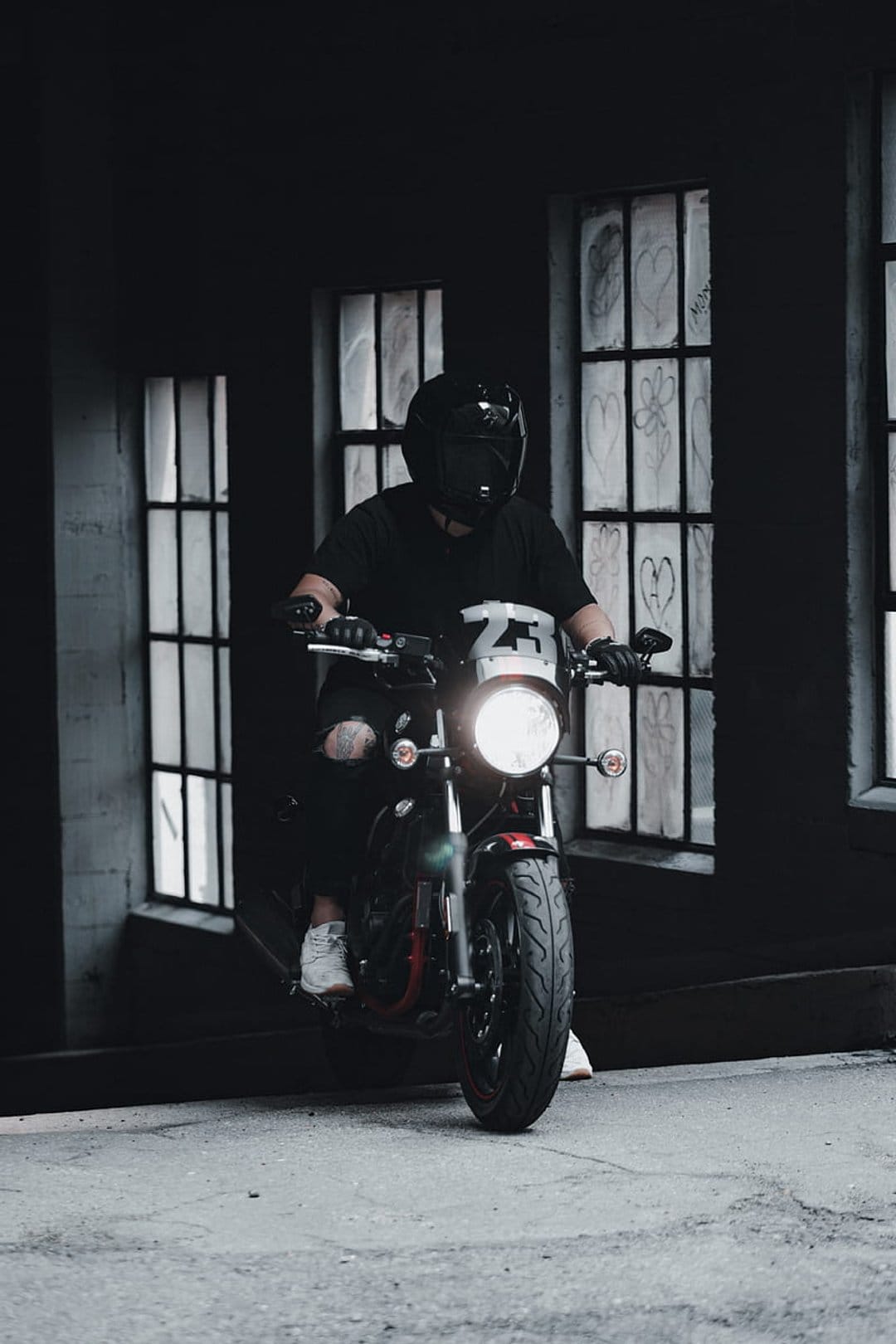Biker in a black helmet sits on a motorcycle with a glowing headlight against a graffiti-covered window backdrop.
