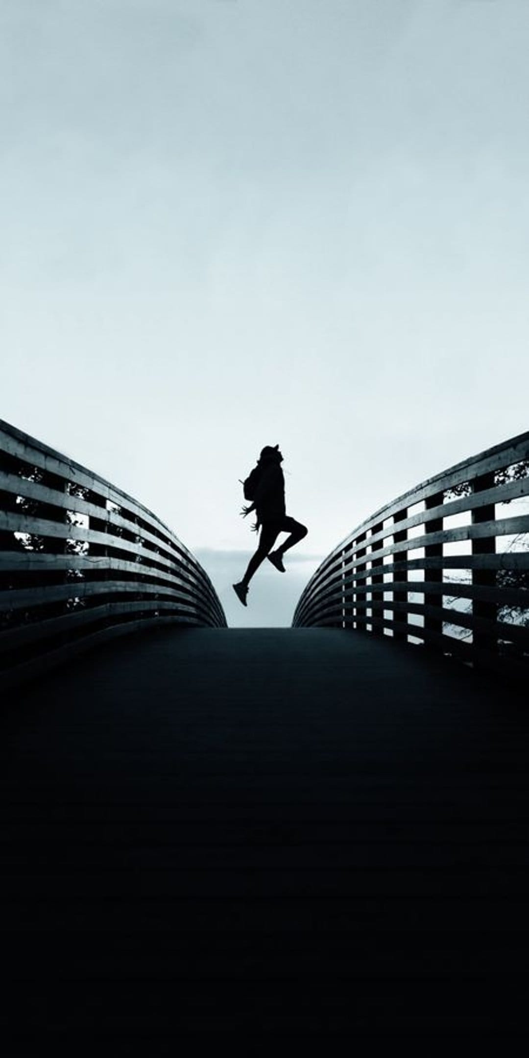 Athletic person in silhouette leaps mid-air between dark bridge railings against a bright minimalist sky.