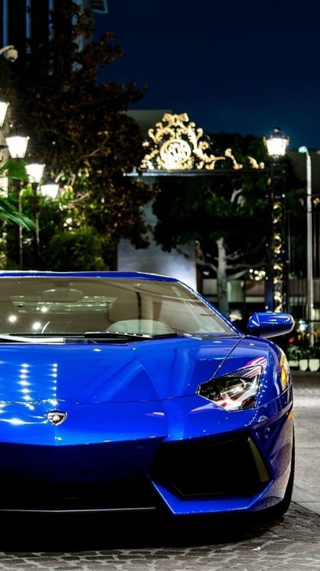 Blue Lamborghini Aventador sports car with glowing LED headlights parked at night near stone pillars and hedges.