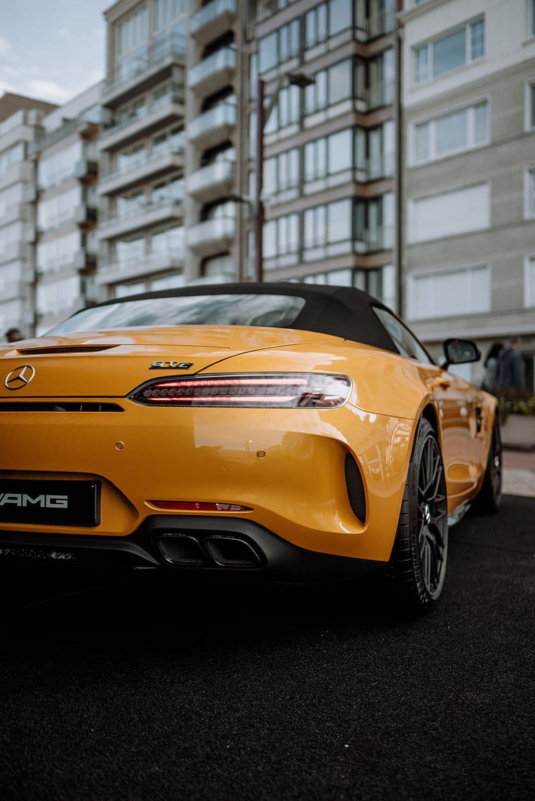 Yellow Mercedes-AMG GT convertible parked on dark pavement behind modern coastal apartments with glass balconies.