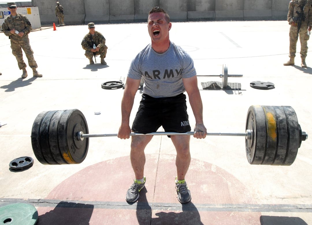 Soldier in an olive green t-shirt performs a heavy deadlift with a loaded barbell in an outdoor training area.