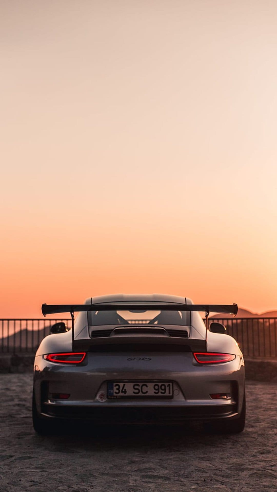 Porsche GT3 RS rear view showing a large wing and red taillights against a sunset sky in vertical orientation.