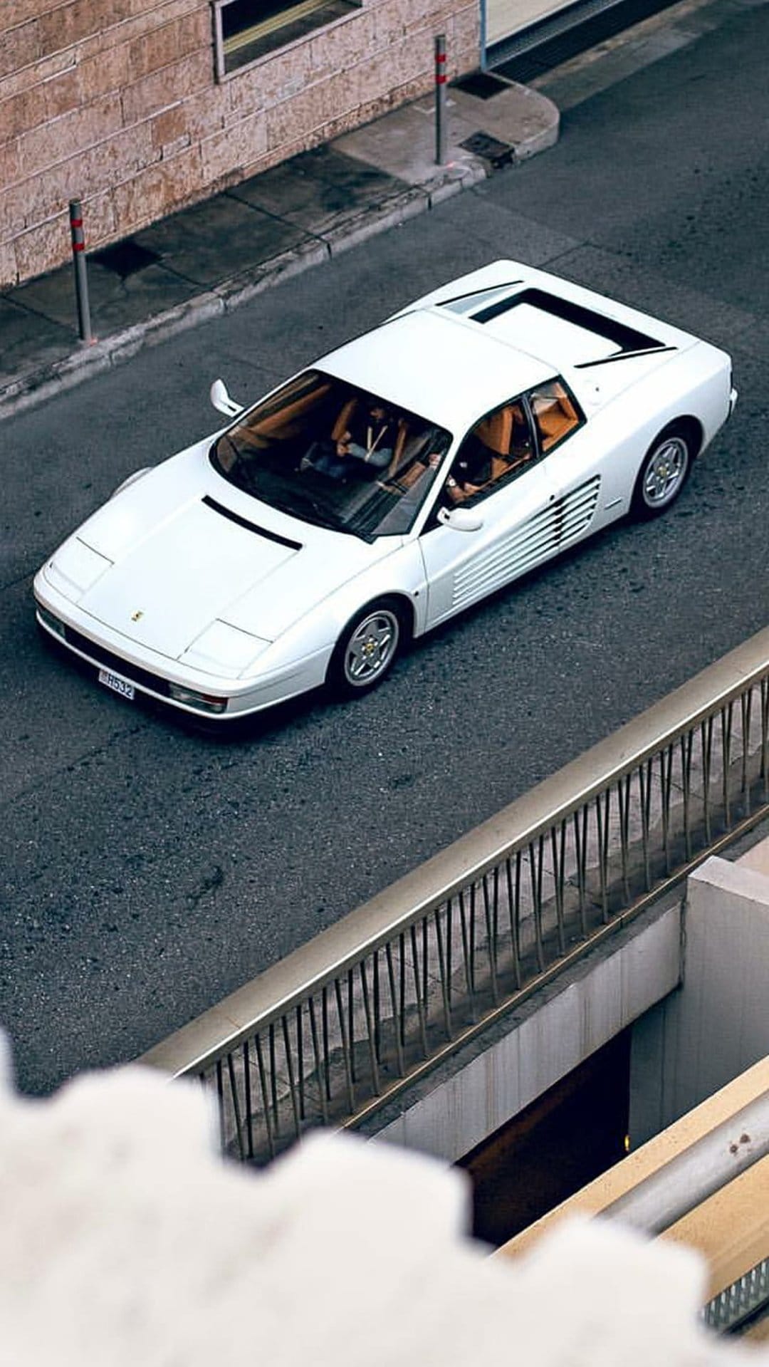 White Ferrari Testarossa driving on dark asphalt seen from an overhead view near a brick building and railing.