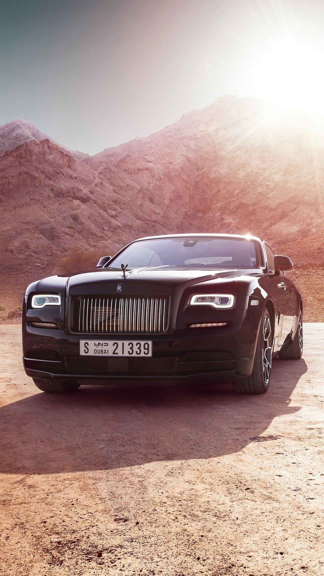 Black Rolls-Royce Wraith in a desert with rugged mountains under intense sunlight and golden hour glow.