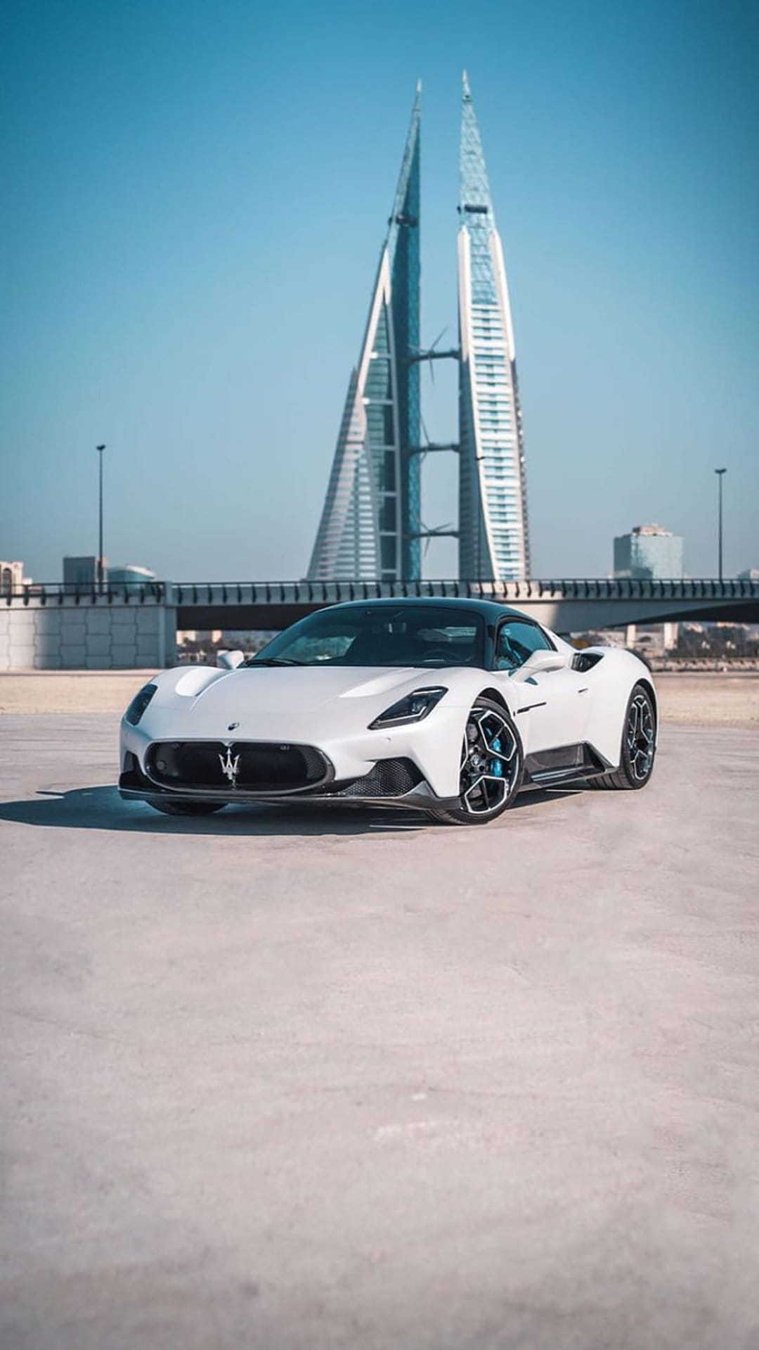 White Maserati MC20 supercar parked in front of the Bahrain World Trade Center skyscrapers under a clear blue sky.
