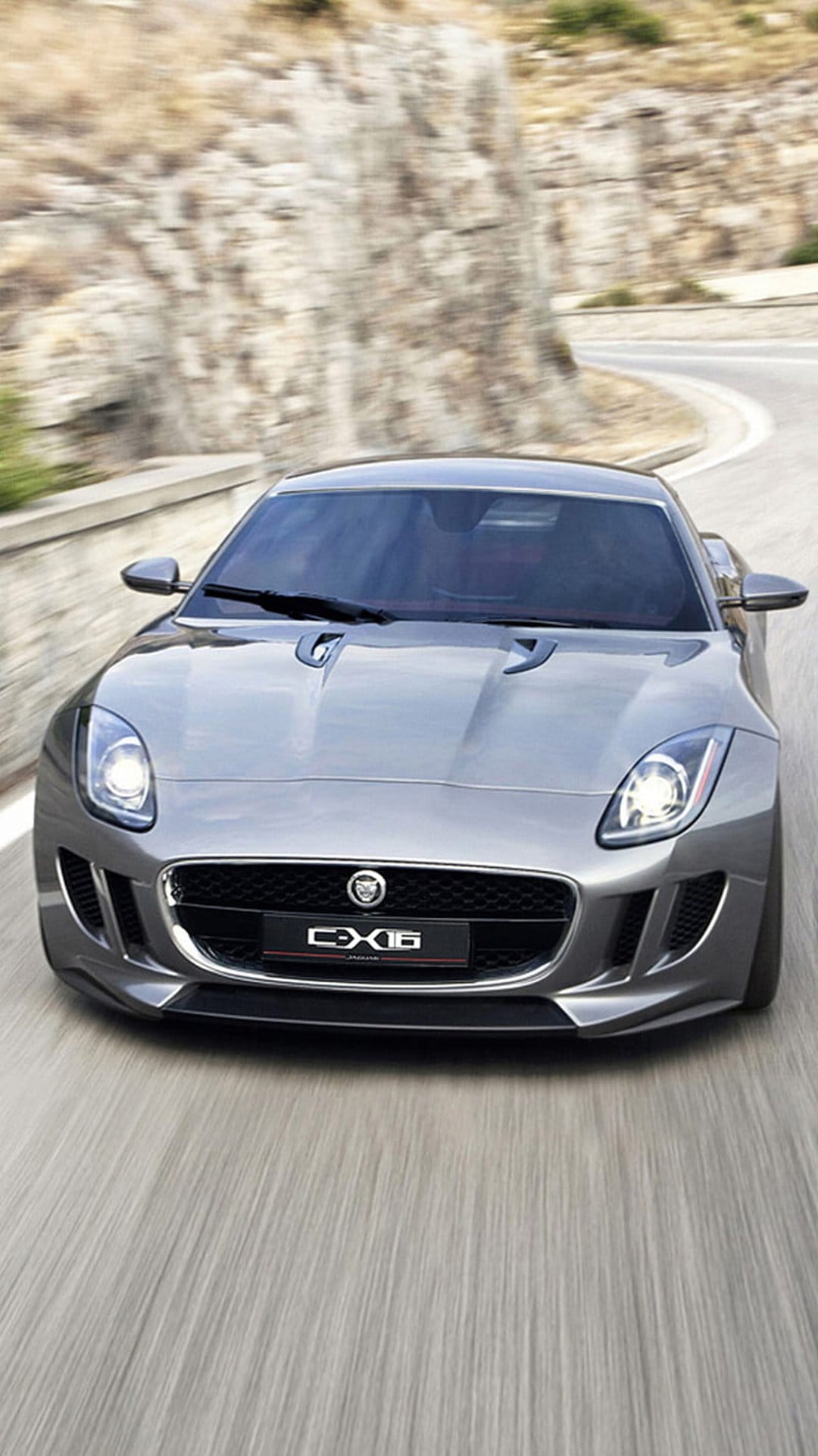 Jaguar CX-16 silver sports car with glowing headlights drives on a paved road next to a rocky mountain hillside.