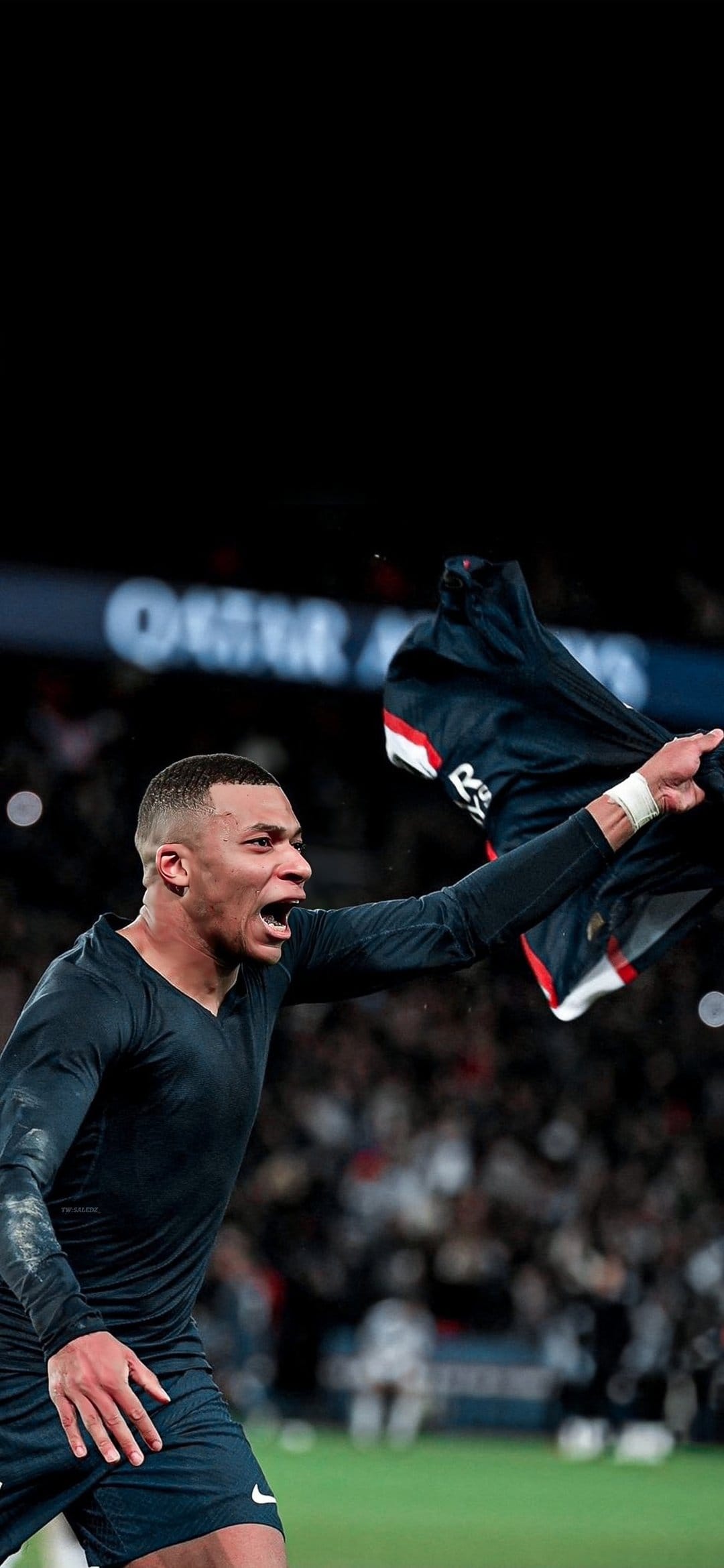 Kylian Mbappe shouts in celebration while wearing a dark PSG jersey inside a stadium with blurred spectators.