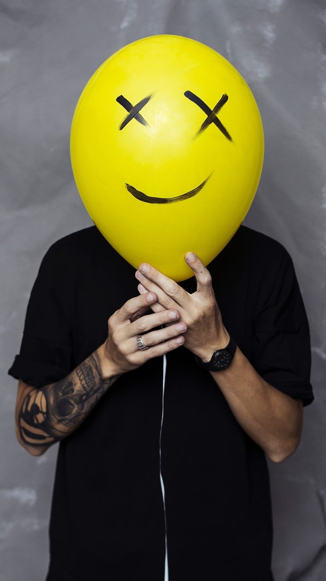 Person with a yellow dead-smiley balloon over their face gestures forward with a tattooed arm and silver watch.