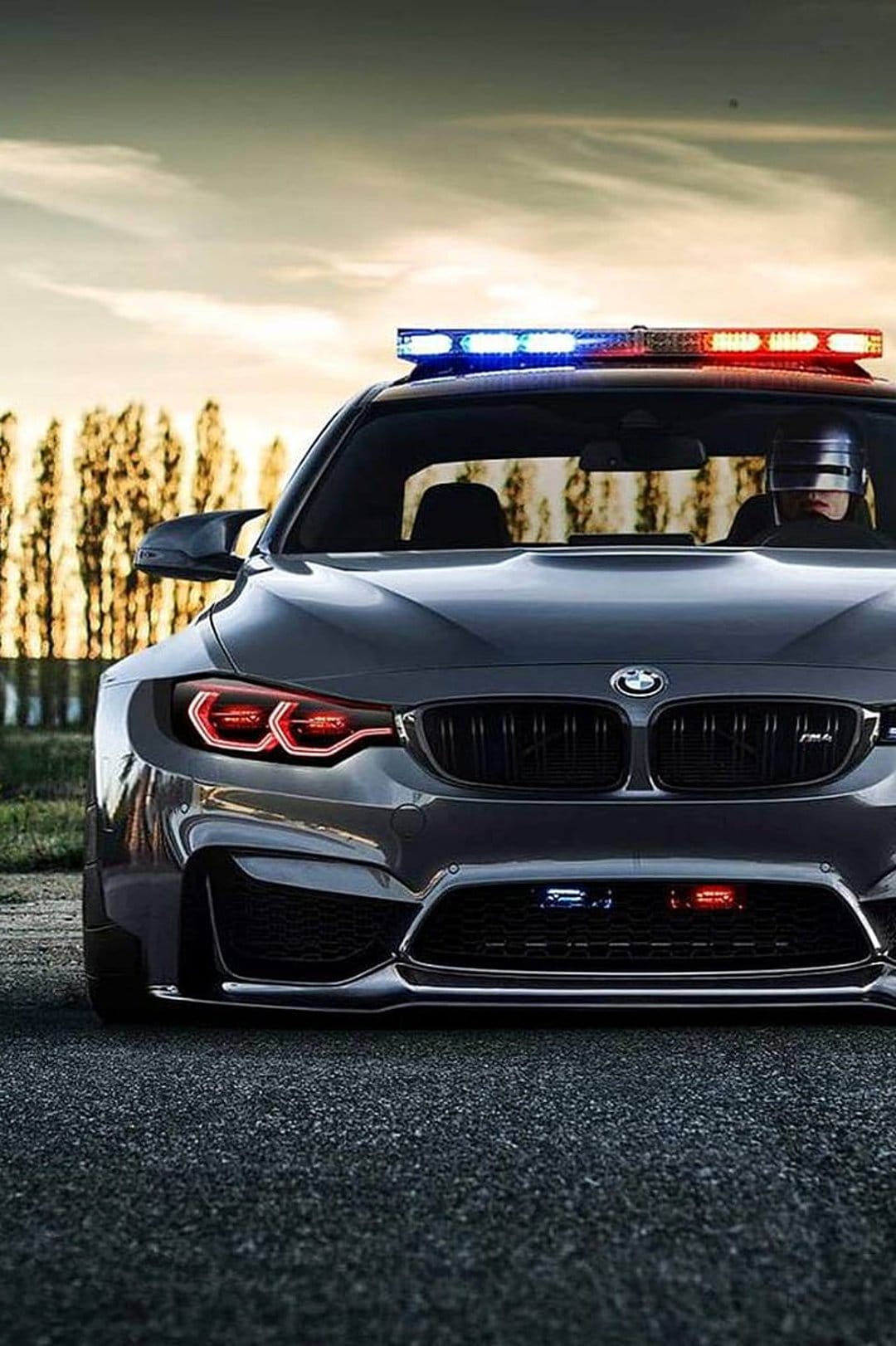 RoboCop wears silver armor while driving a gray BMW M4 police car with flashing blue and red emergency lights.