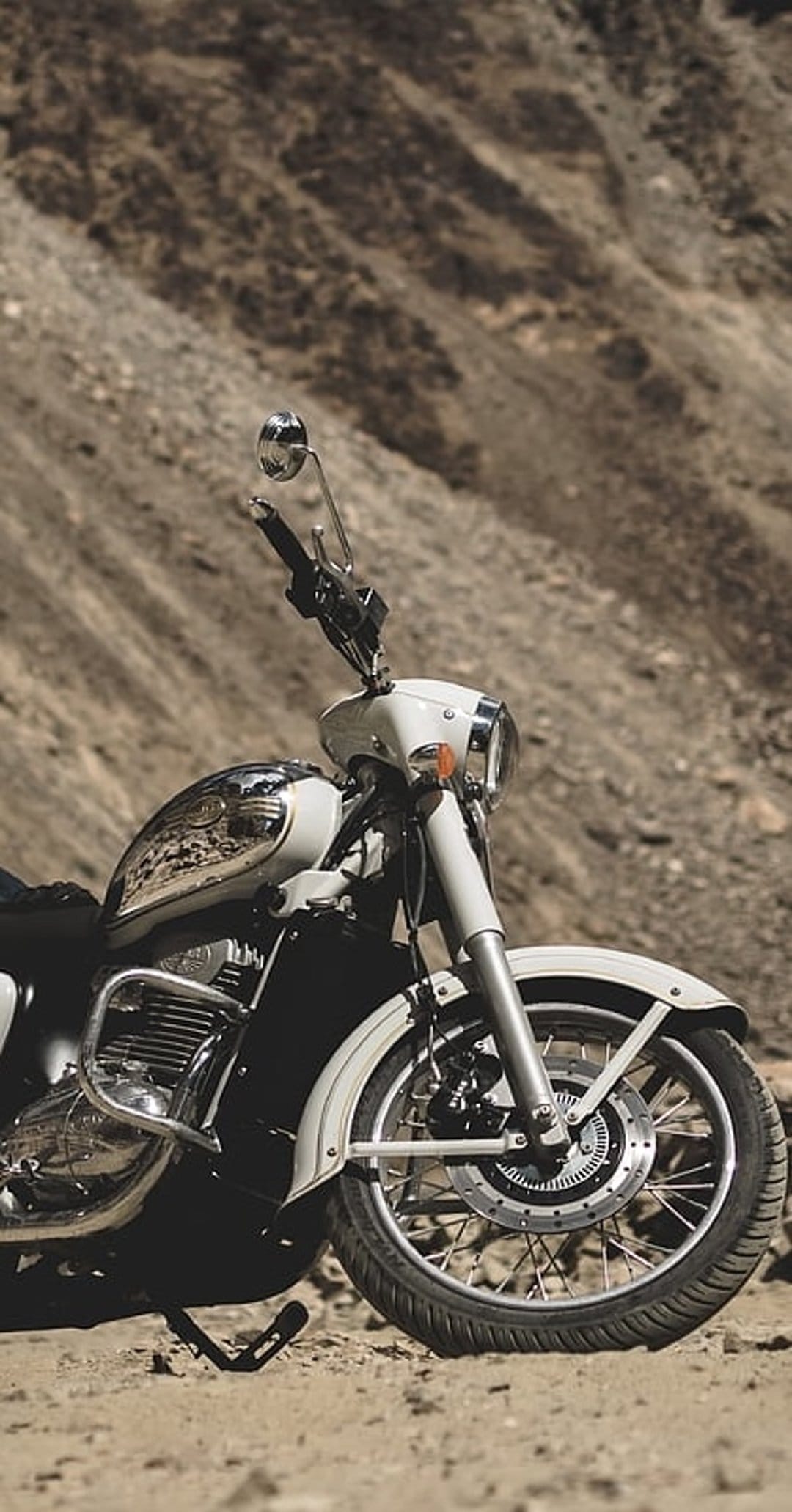 White vintage motorcycle with a chrome tank parked on a rocky trail against a blurred mountain range backdrop.