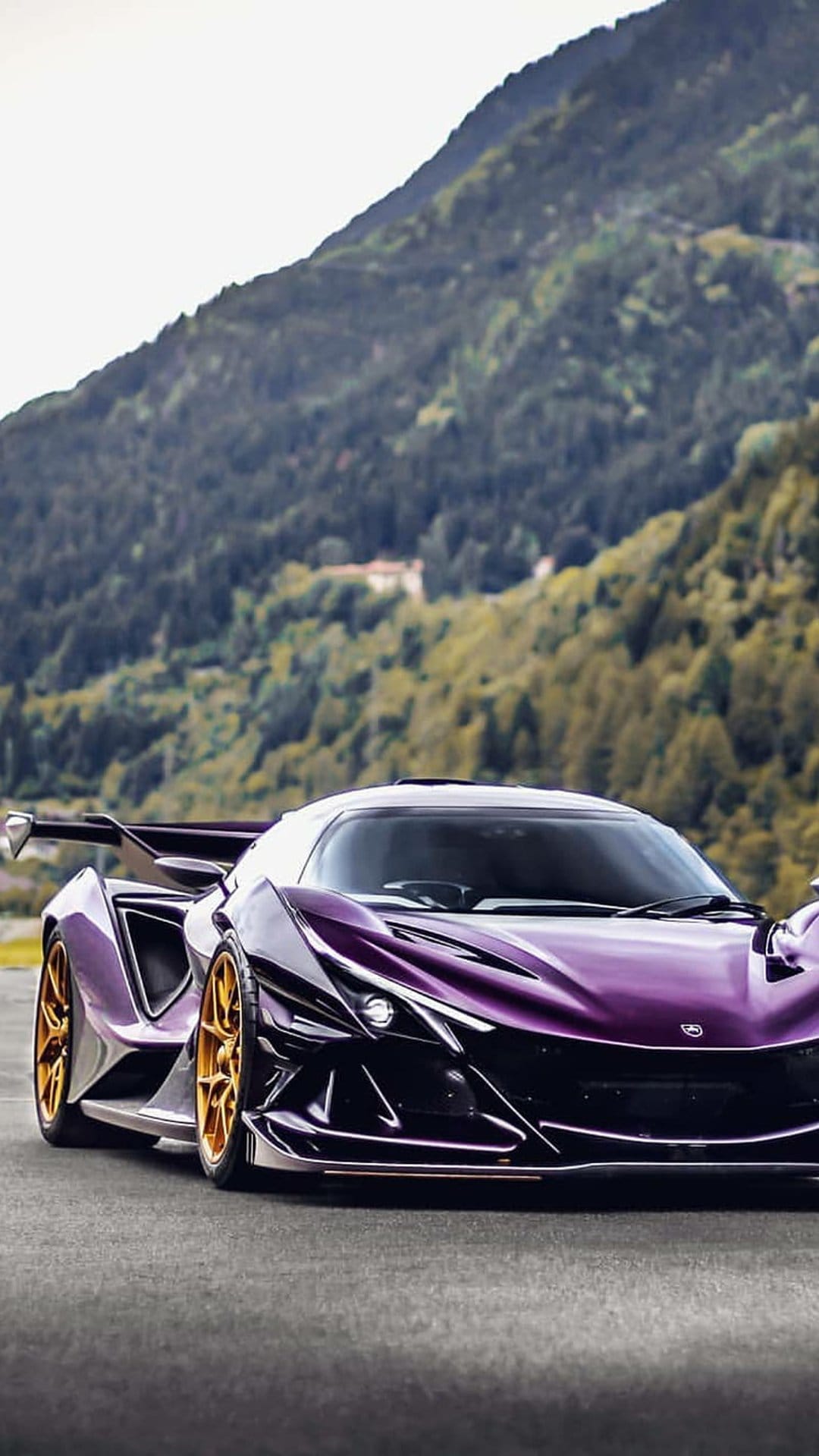 Purple Apollo hypercar with gold rims drives on a paved mountain road surrounded by a dense green pine forest.