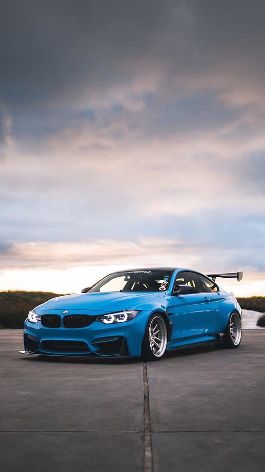 Blue BMW M4 Coupe with a large race spoiler parked on concrete under a cloudy, overcast coastal sky.