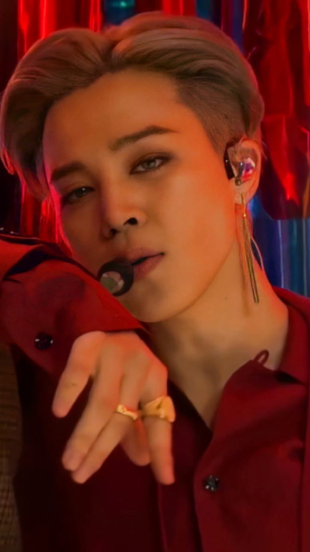 Jimin from BTS holds a microphone wearing a red shirt and silver hair during a dramatic stage performance.