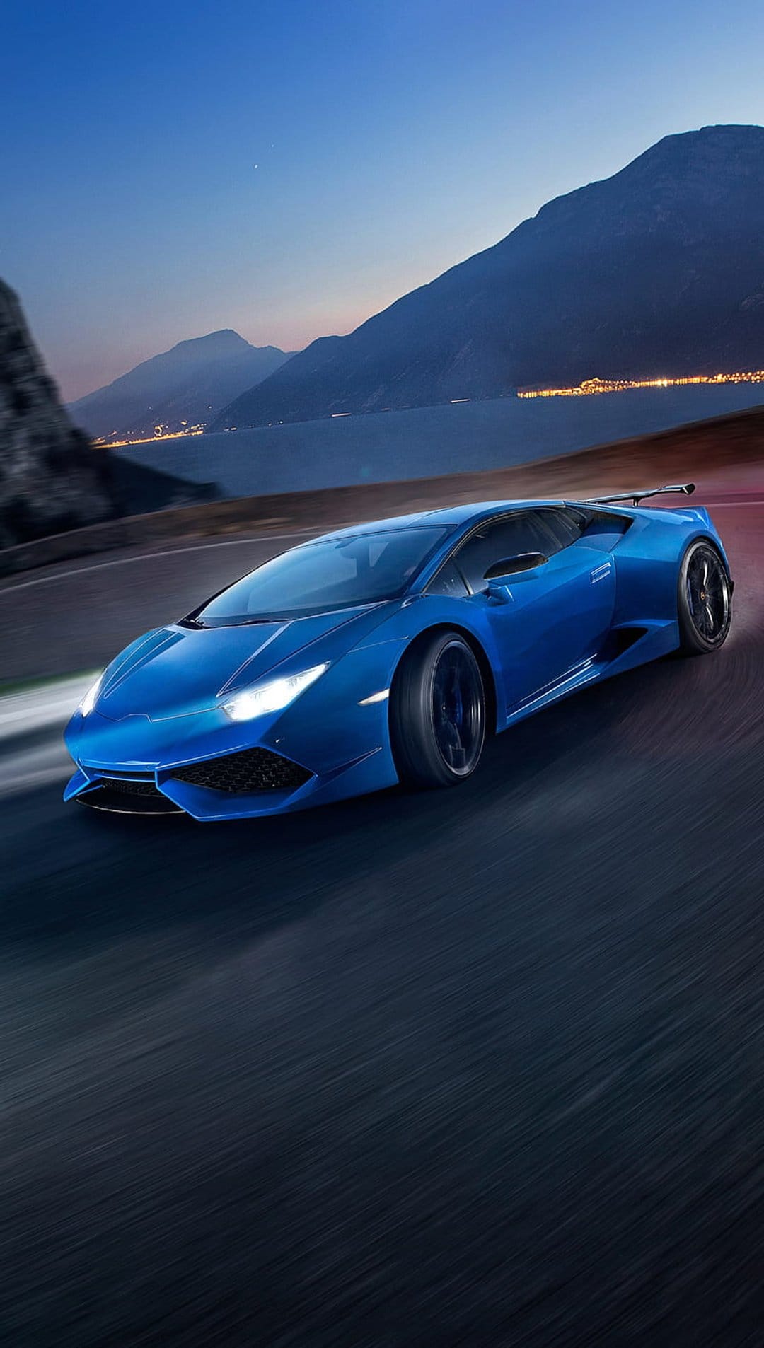 Blue Lamborghini supercar driving on a winding mountain road at dusk with city lights and water in the background.